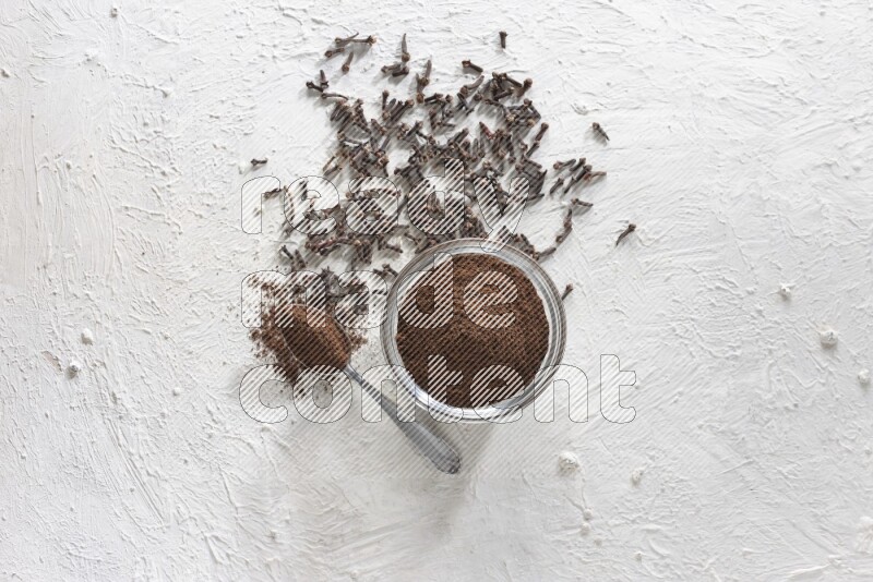 A glass bowl and a metal spoon full of cloves powder with cloves grains spread on a textured white flooring