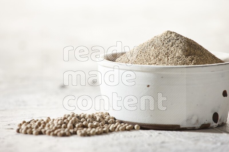 white pottery bowl full of white pepper powder with pepper beads on textured white flooring