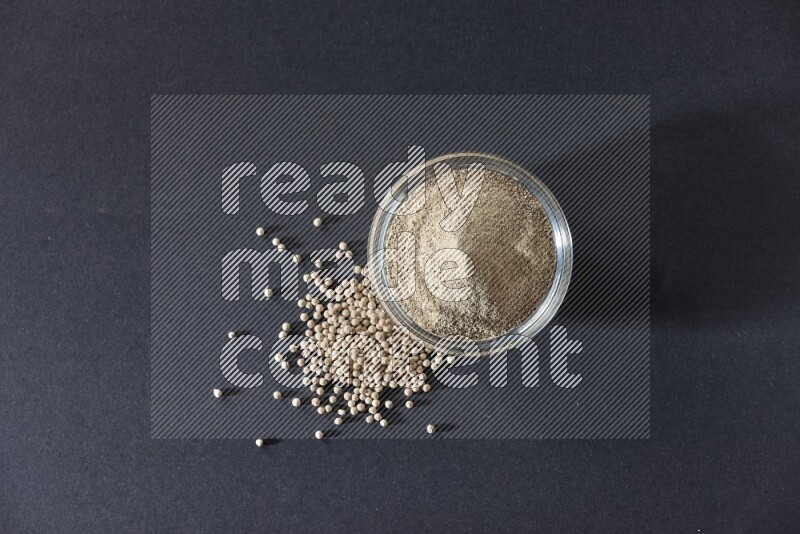 A glass bowl full of white pepper powder with white pepper beads on black flooring