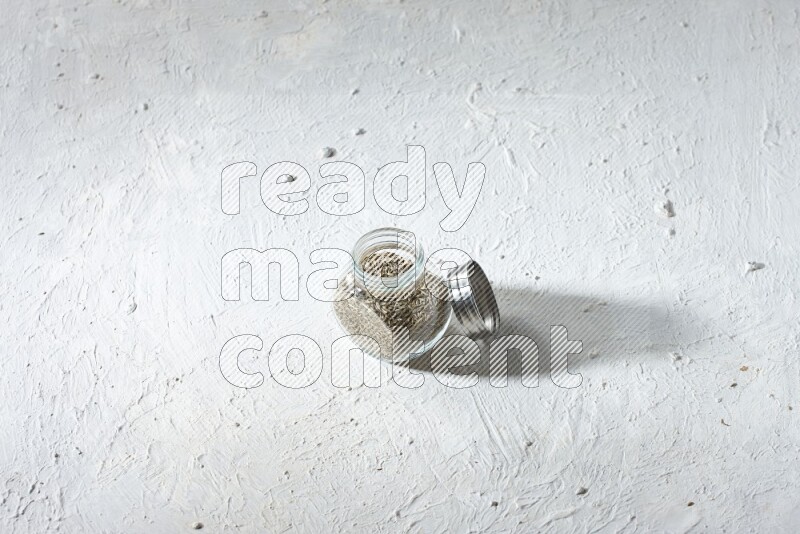 A glass spice jar full of cumin seeds on textured white flooring