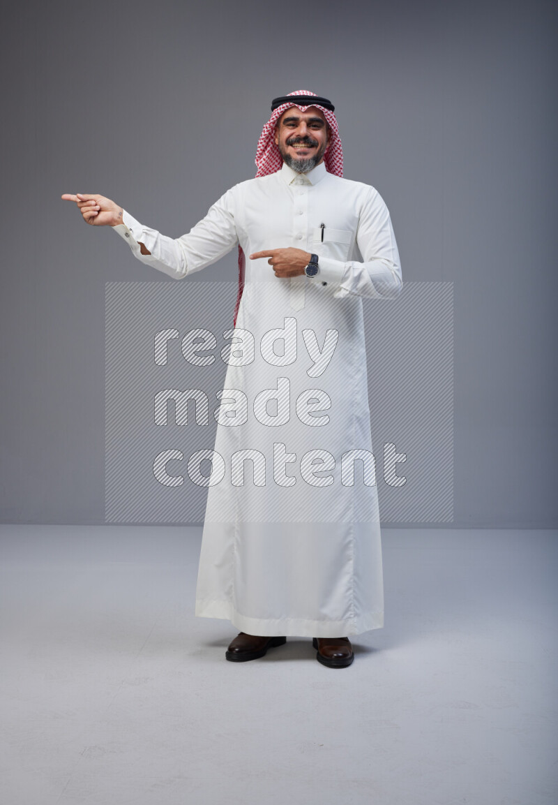 Saudi man Wearing Thob and red white Shomag standing interacting with the camera on Gray background