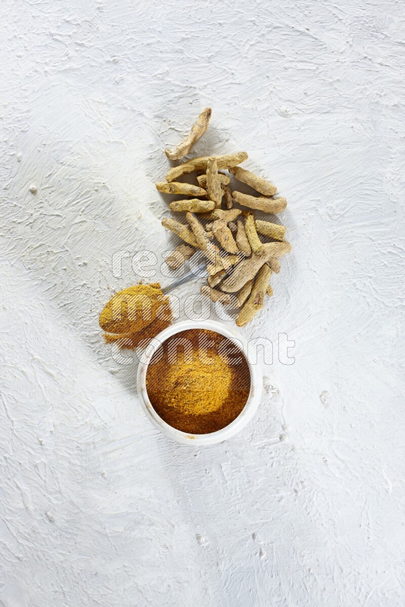 A beige pottery bowl and metal spoon full of turmeric powder and dried turmeric fingers next of them on textured white flooring
