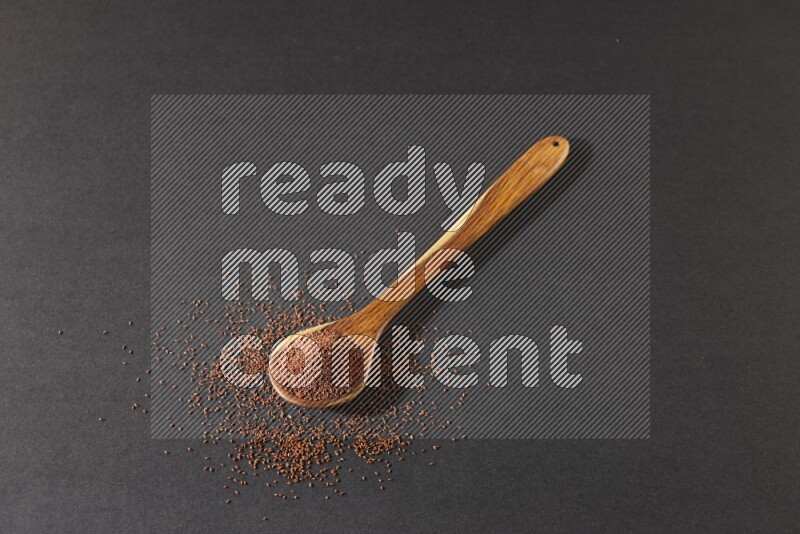 A wooden ladle full of garden cress seeds and seeds spread beside it on a black flooring