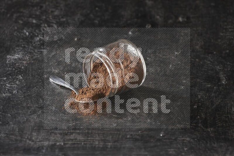 A flipped glass jar and metal spoon full of cloves powder with cloves spread on a textured black flooring