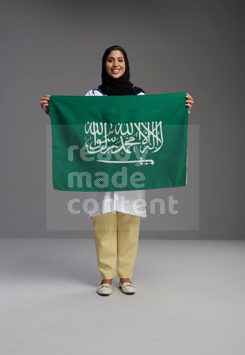 Saudi woman wearing lab coat with stethoscope standing holding Saudi flag on Gray background