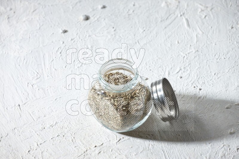 A glass spice jar full of cumin seeds on textured white flooring