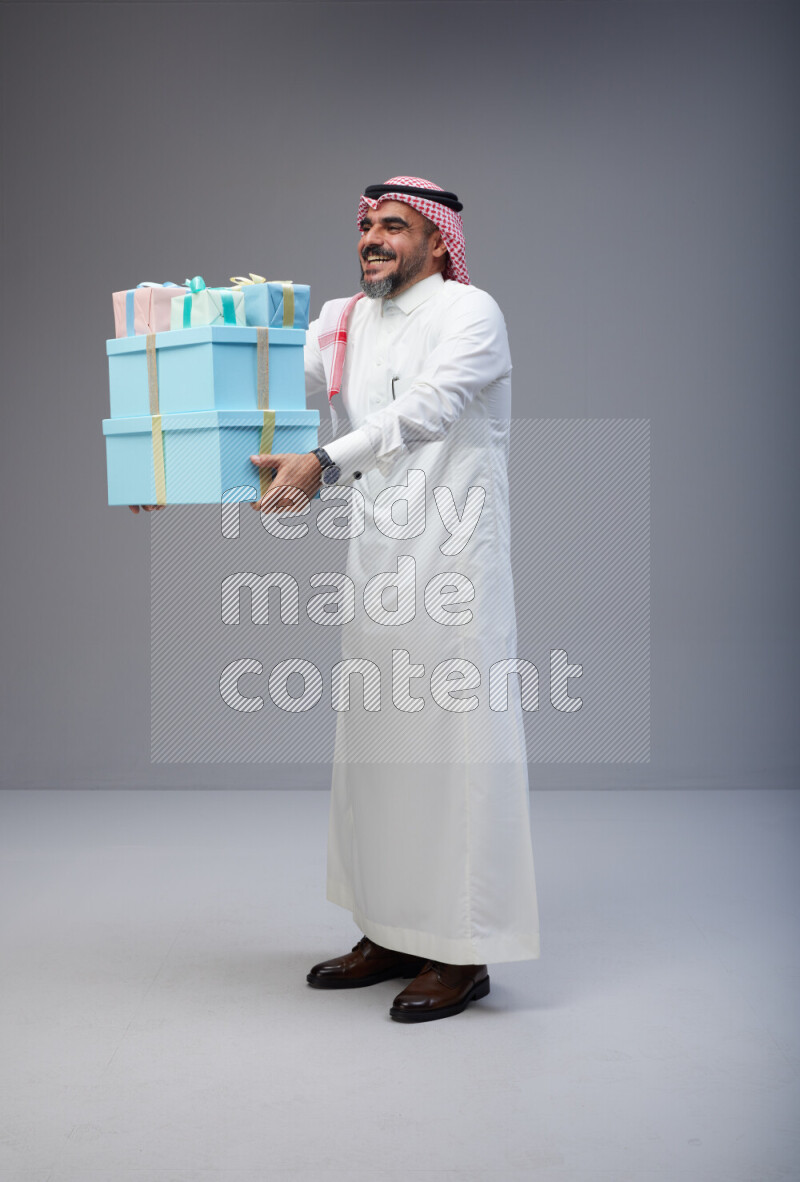 Saudi man Wearing Thob and red Shomag standing holding gift box on Gray background