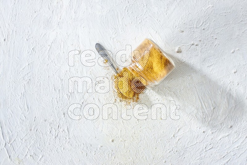 A flipped glass spice jar and a metal spoon full of turmeric powder and powder spilled out of it on textured white flooring