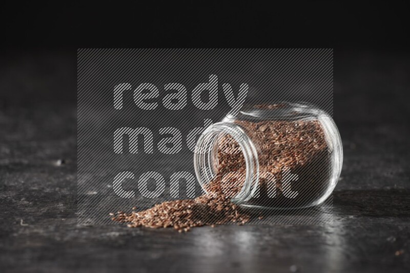 A glass spice jar full of flaxseeds flipped and seeds spread out on a textured black flooring