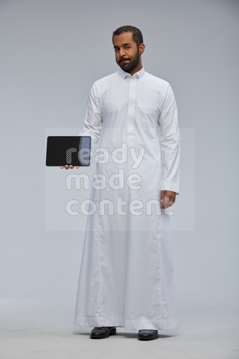 Saudi man wearing Thob standing showing tablet to camera on Gray background