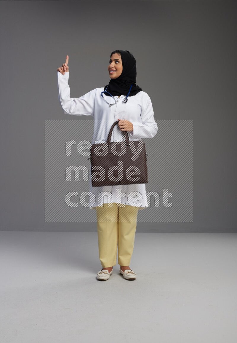 Saudi woman wearing lab coat with stethoscope standing holding bag on Gray background