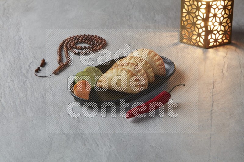 Four fried sambosas in an oval shaped black plate, beside a cut cheese sambosa, a brown misbaha and a golden lantern on a gray background