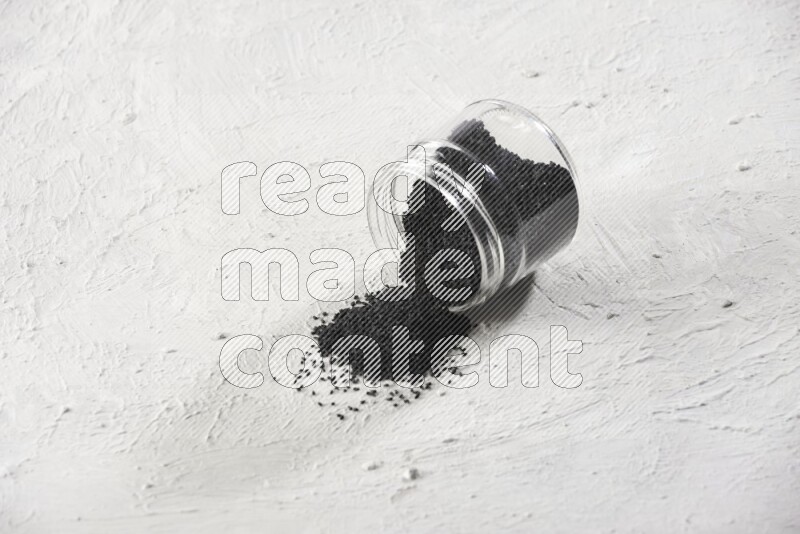 A glass jar full of black seeds and the jar flipped and seeds spread on a textured white flooring