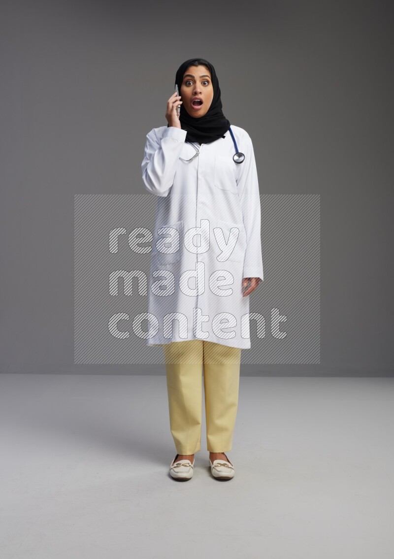 Saudi woman wearing lab coat with stethoscope standing talking on phone on Gray background