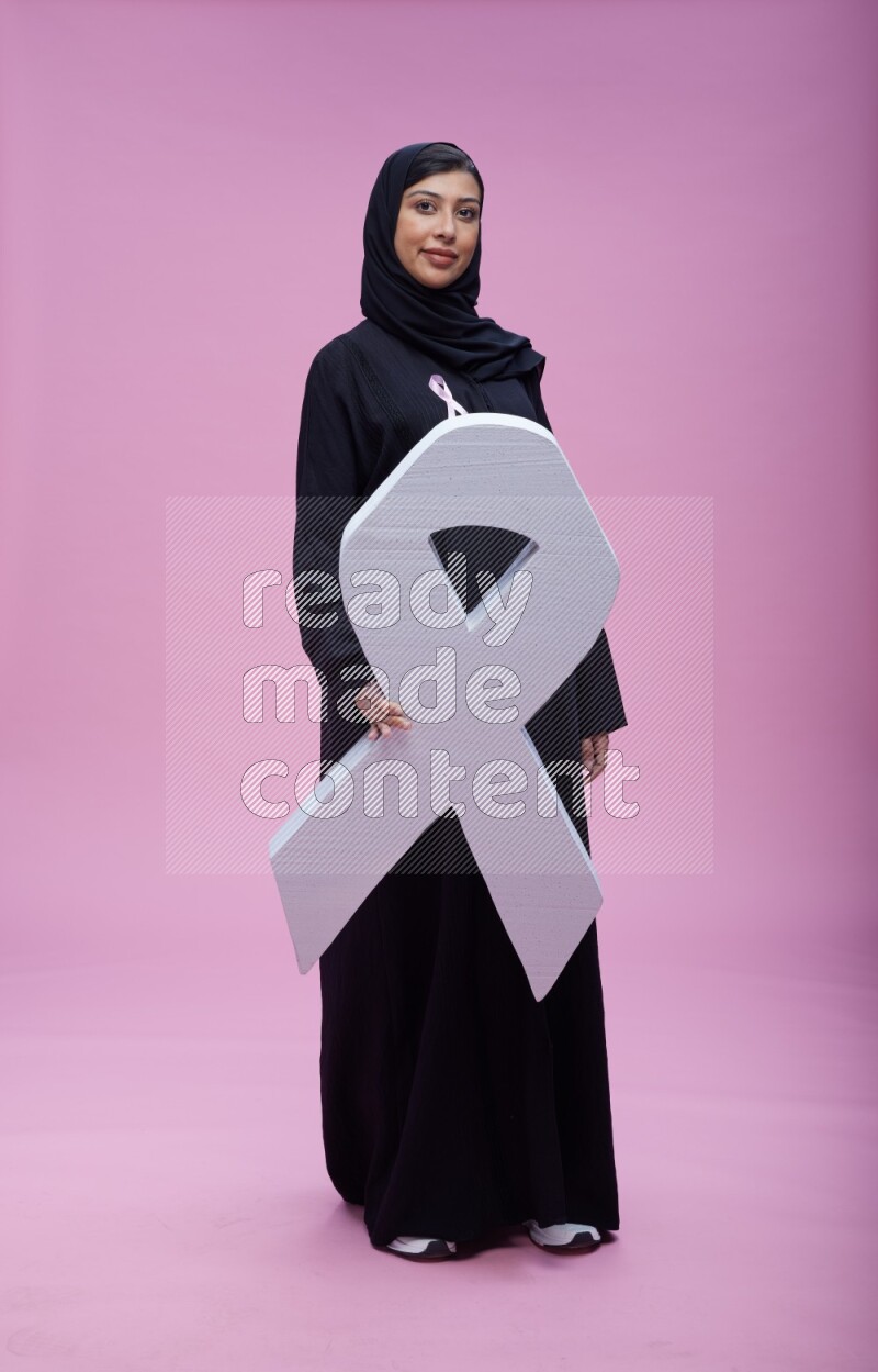 Saudi woman wearing pink ribbon on abaya standing holding awareness ribbon on plain pink background