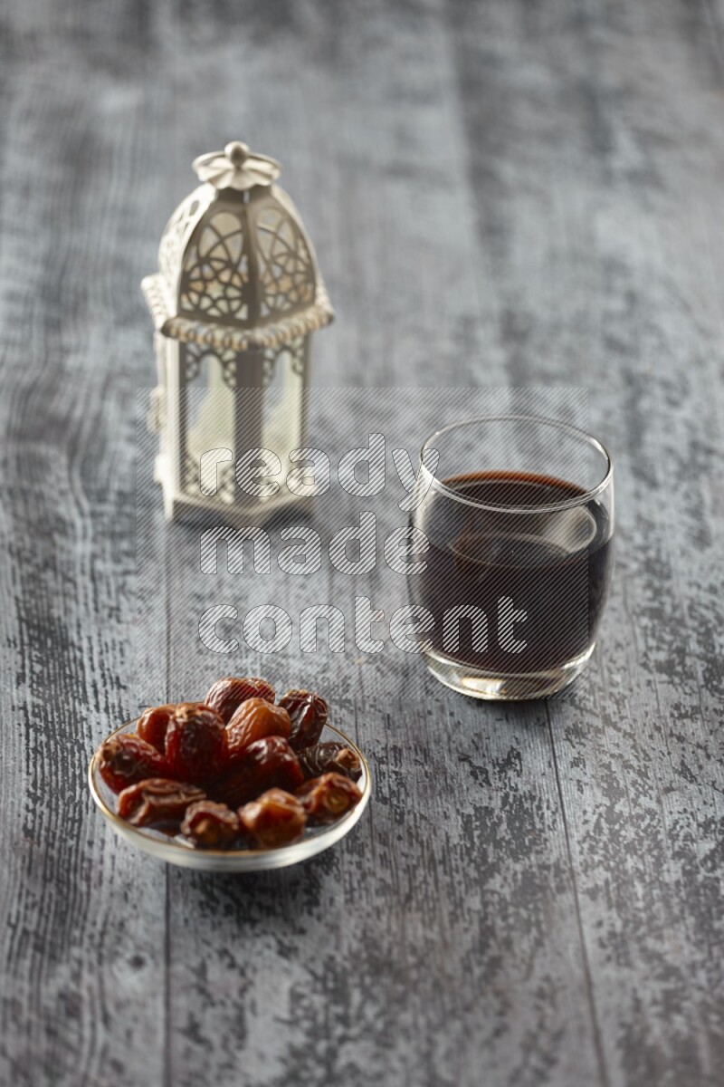 A white lantern with different drinks, dates, nuts, prayer beads and quran on grey wooden background