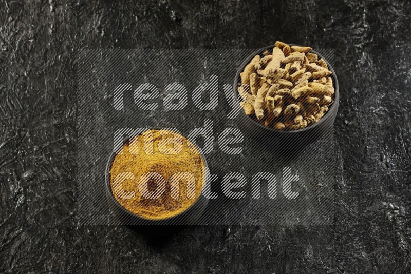 2 pottery black bowls, one full of turmeric powder and the other full of dried turmeric whole fingers on a textured black flooring