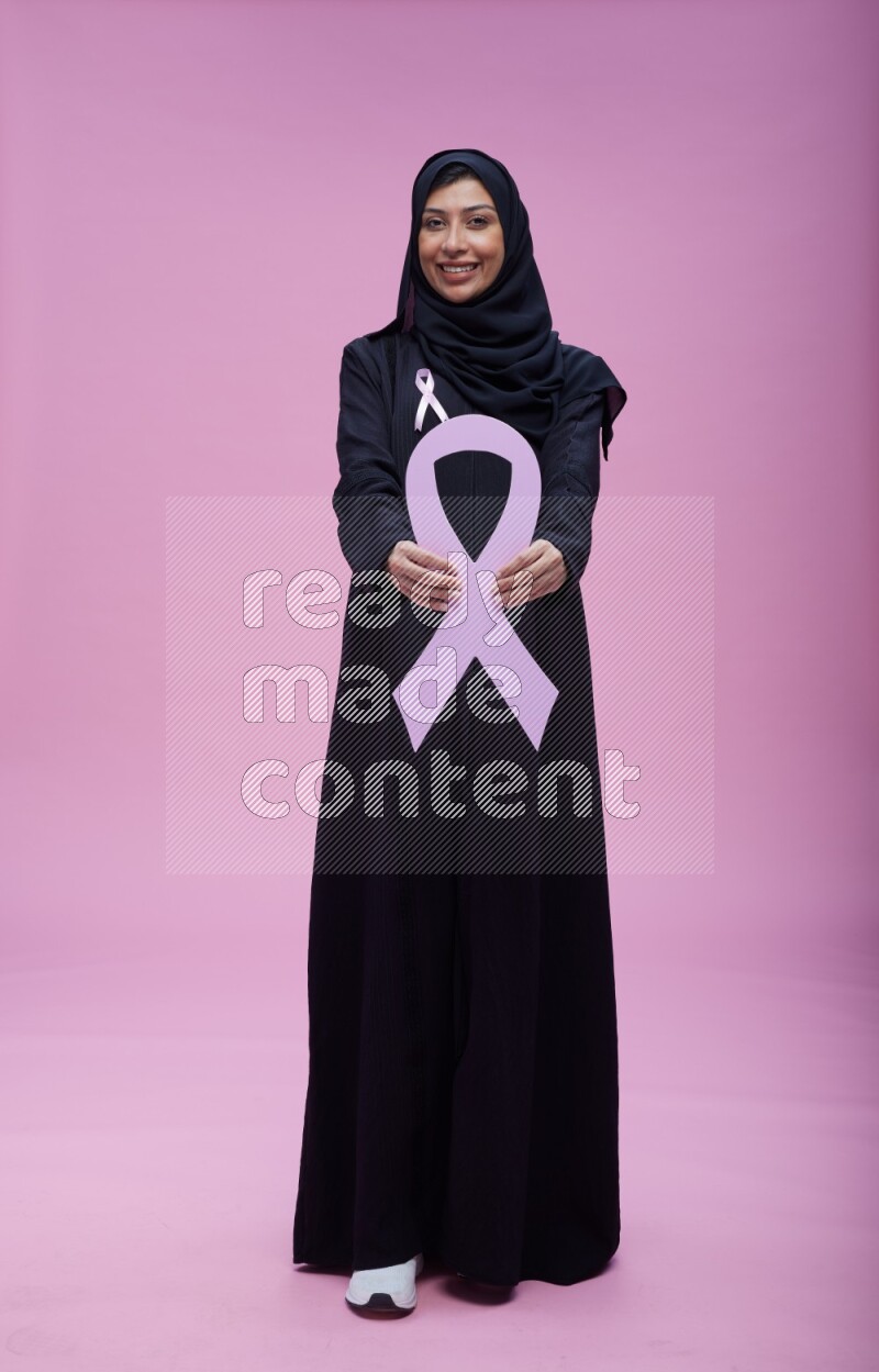 Saudi woman wearing pink ribbon on abaya standing holding awareness ribbon on plain pink background