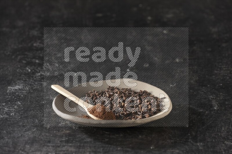 A Pottery plate full of cloves and a wooden spoon full of cloves powder on it on a textured black background