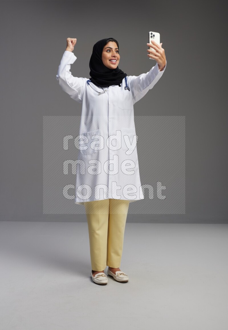 Saudi woman wearing lab coat with stethoscope standing taking selfie on Gray background