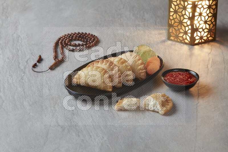 Four fried sambosas in an oval shaped black plate, beside a cut cheese sambosa, a brown misbaha and a golden lantern on a gray background