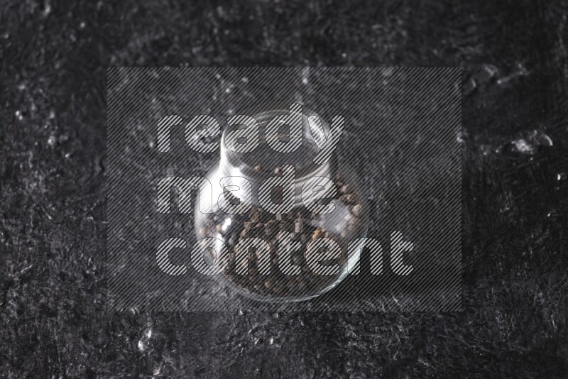 A glass spice jar full of black pepper beads on textured black flooring