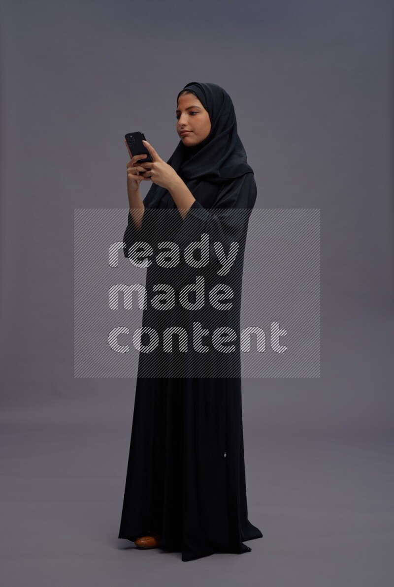 Saudi woman wearing Abaya standing texting on phone on gray background