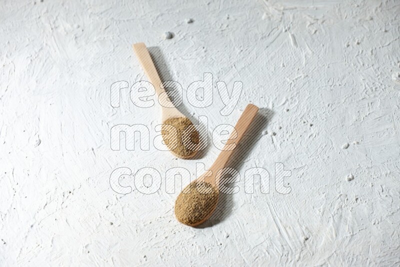 2 Wooden spoons full of cumin powder on a textured white flooring