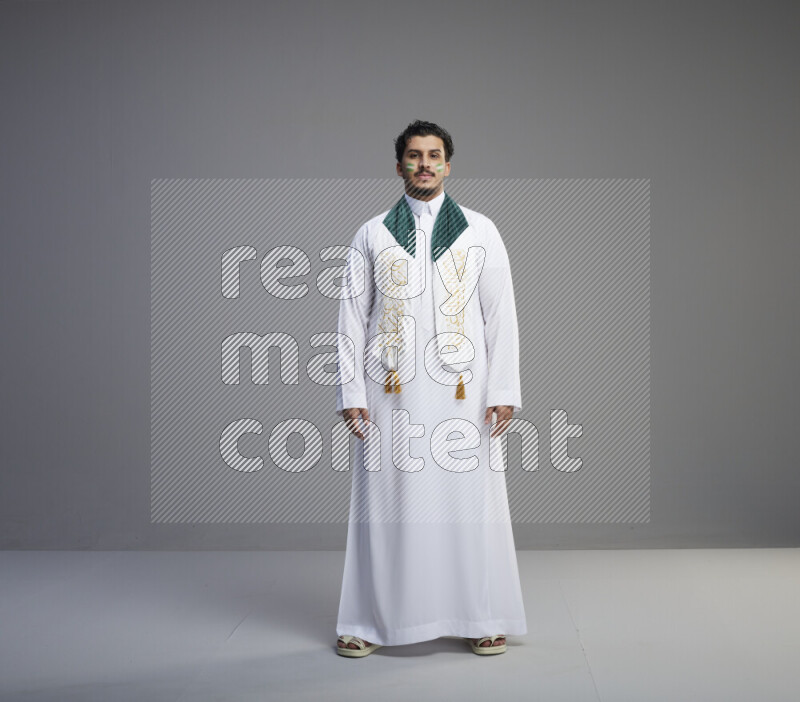 A Saudi man standing wearing thob and Saudi flag scarf with face painting on gray background