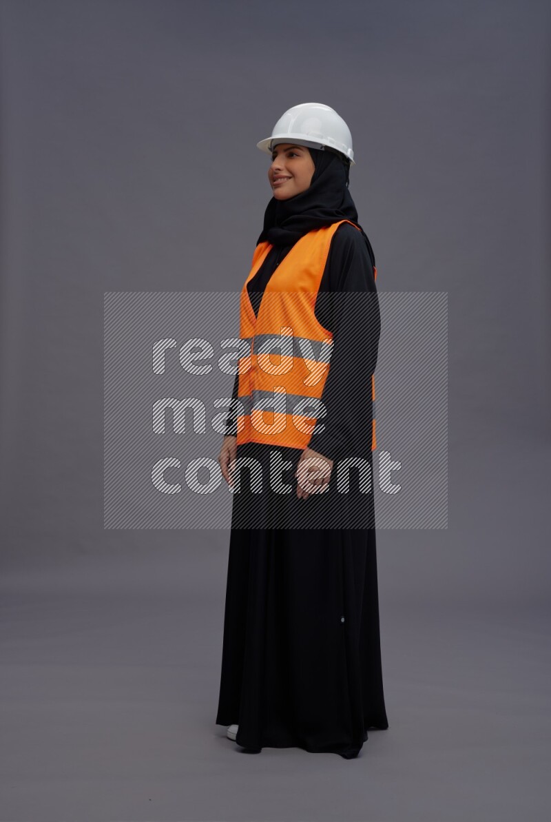 Saudi woman wearing Abaya with engineer vest standing interacting with the camera on gray background