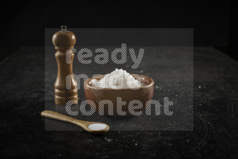 A wooden bowl and spoon filled with white sea salt and wooden grinder beside them on black background