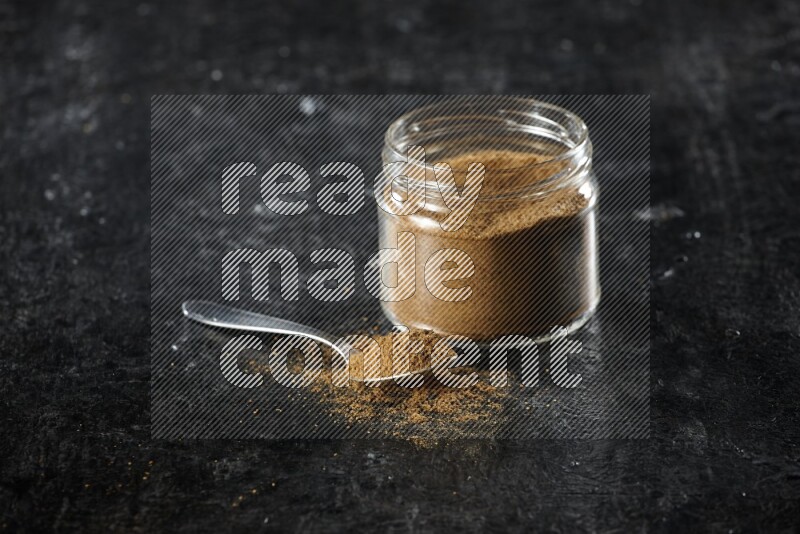 A glass jar and a metal spoon full of cumin powder on a textured black flooring