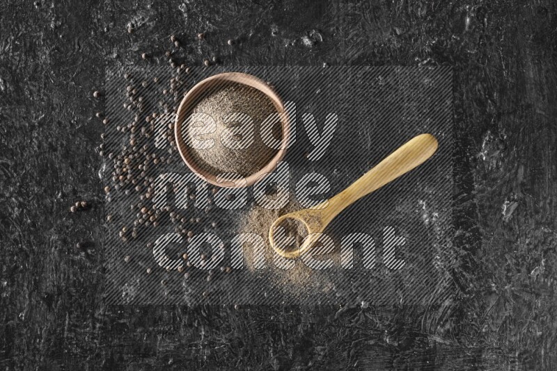 A wooden bowl and a wooden spoon full of black pepper powder with the beads on a textured black flooring