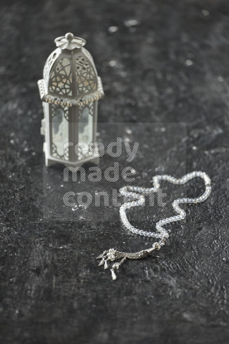 A white lantern with different drinks, dates, nuts, prayer beads and quran on textured black background