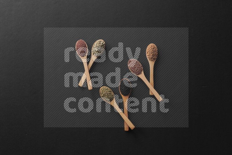wooden spoons filled with white peppers, mustard seeds, black seeds, garden cress, cumin and flax on black flooring