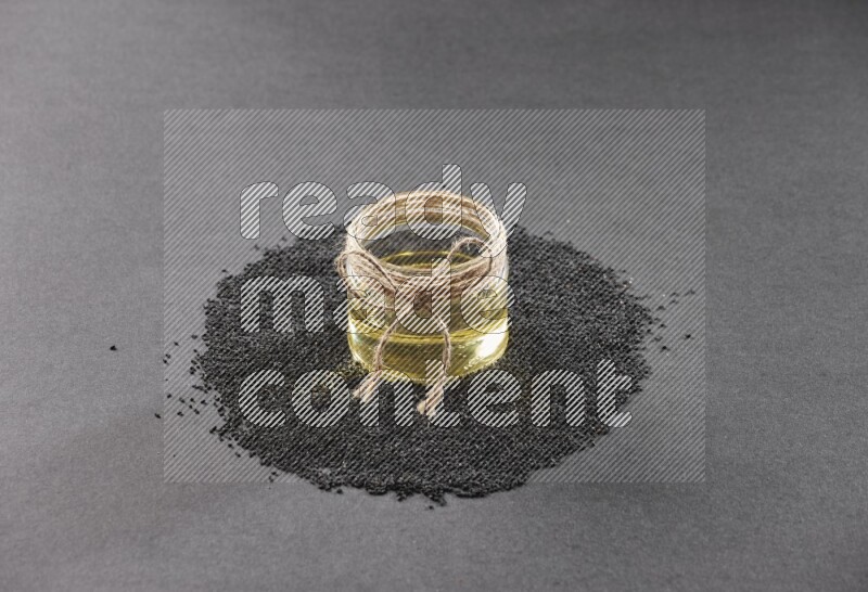 A glass jar full of black seeds oil surrounded by the seeds on a black flooring