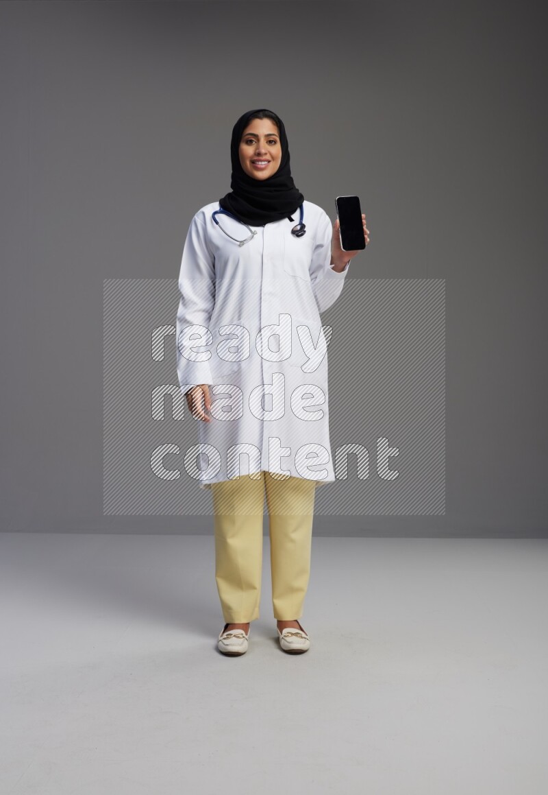Saudi woman wearing lab coat with stethoscope standing showing phone to camera with sign in the back on Gray background