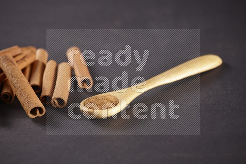 Cinnamon sticks stacked beside a wooden spoon full of cinnamon powder on black background