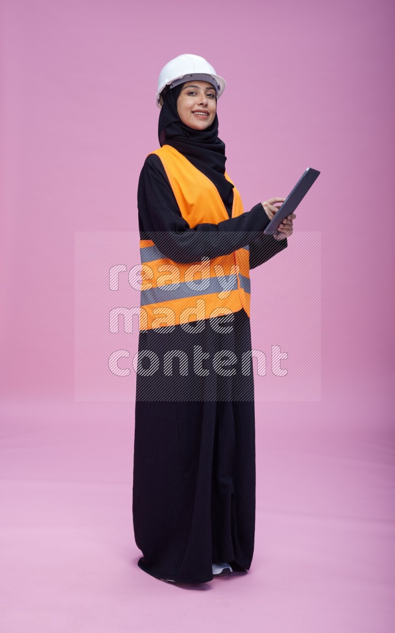 Saudi woman wearing Abaya with engineer vest and helmet standing working on tablet on pink background