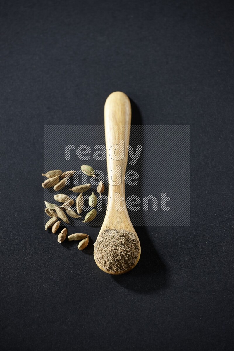 A wooden spoon full of cardamom powder and cardamom seeds beside it on black flooring