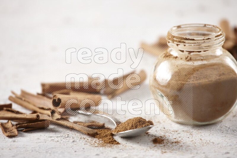 Herbal glass jar full cinnamon powder and a metal spoon surrounded by cinnamon sticks on a white background