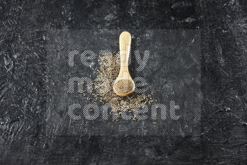 A wooden spoon full of cumin powder and cumin seeds spreaded on textured black flooring