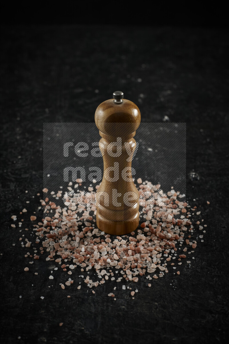 A wooden grinder standing upright and surrounded by coarse pink himalayan salt on black background