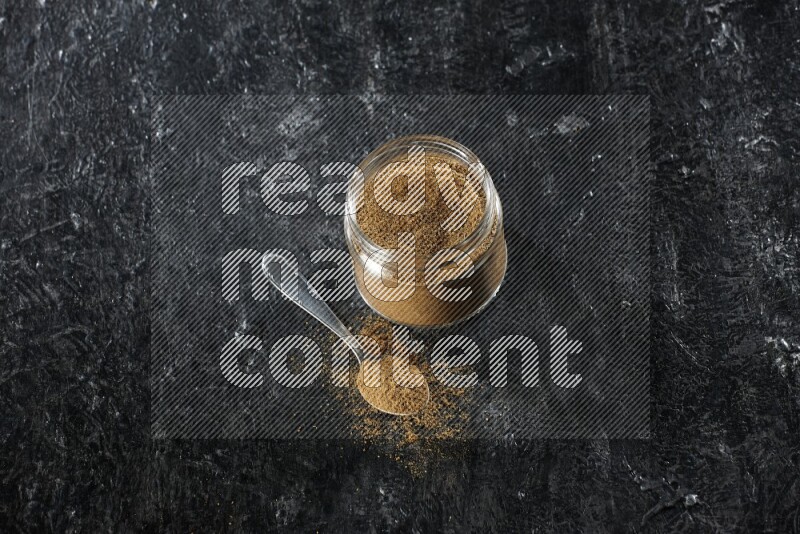 A glass jar and a metal spoon full of cumin powder on a textured black flooring