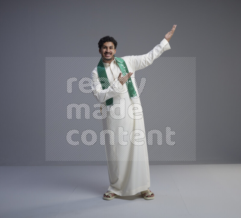 A Saudi man standing wearing thob and Saudi flag scarf on gray background