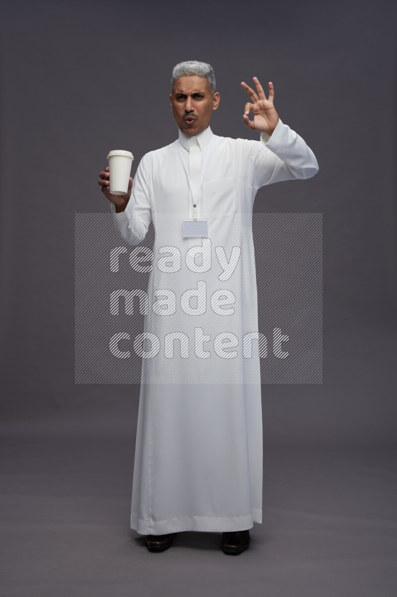 Saudi man wearing thob with neck strap employee badge standing holding paper cup on gray background