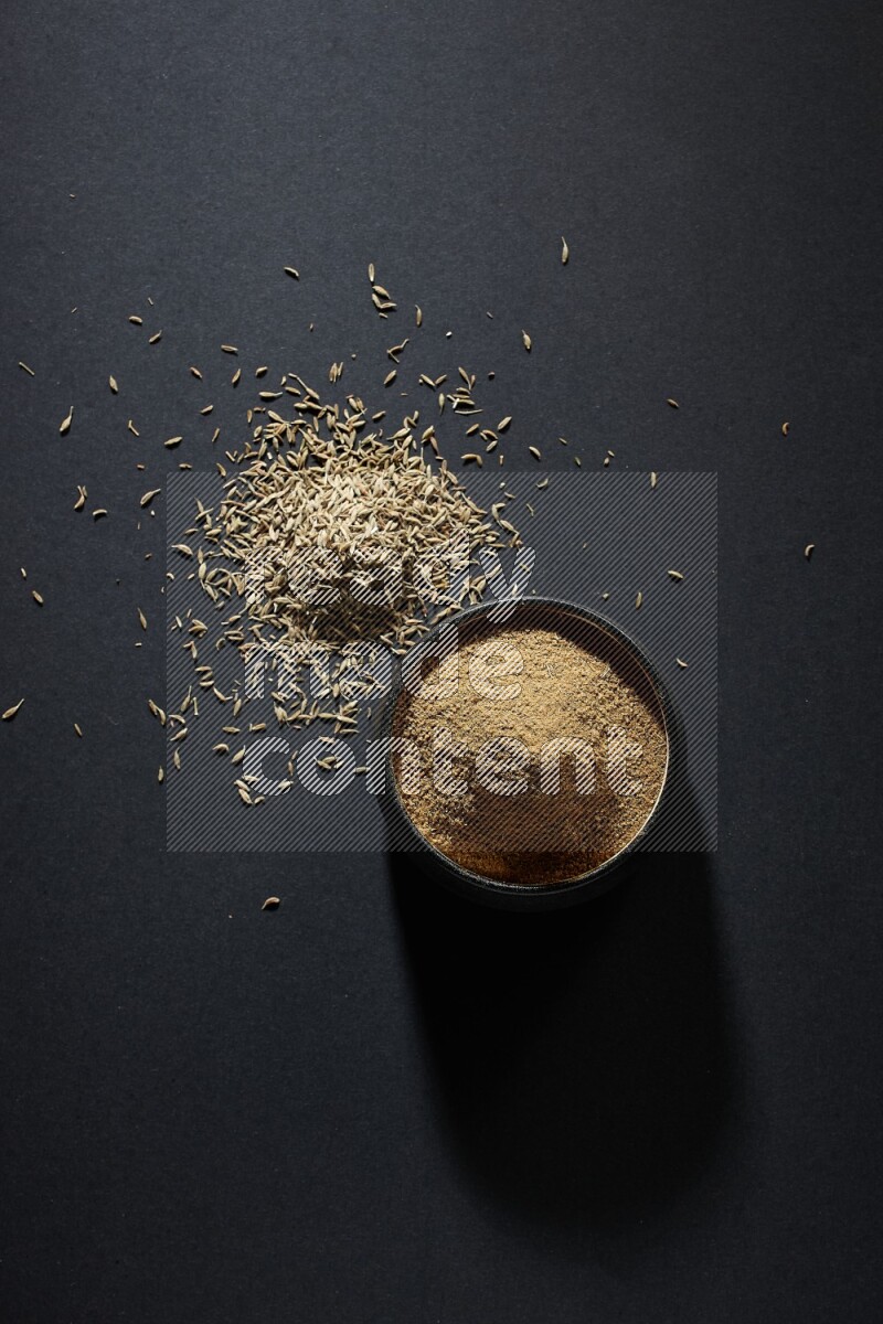 A black pottery bowl full of cumin powder and spreaded cumin seeds on a black flooring