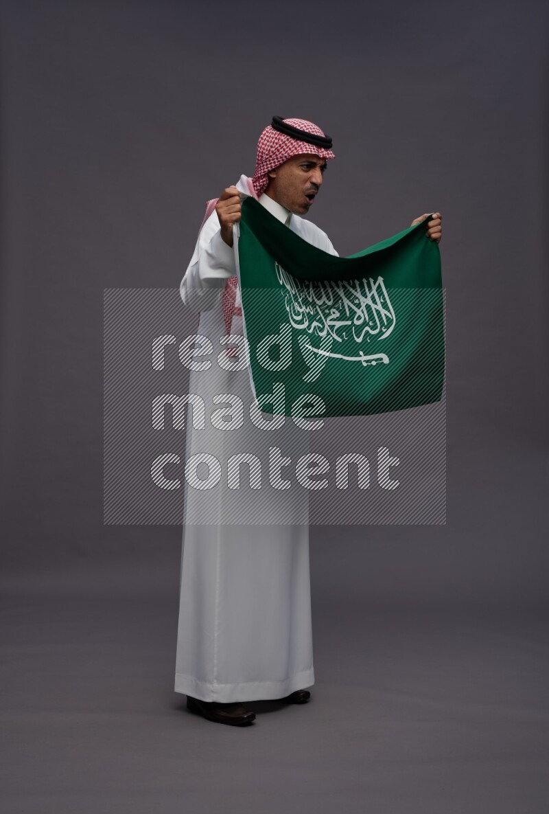 Saudi man wearing thob and shomag standing holding Saudi flag on gray background