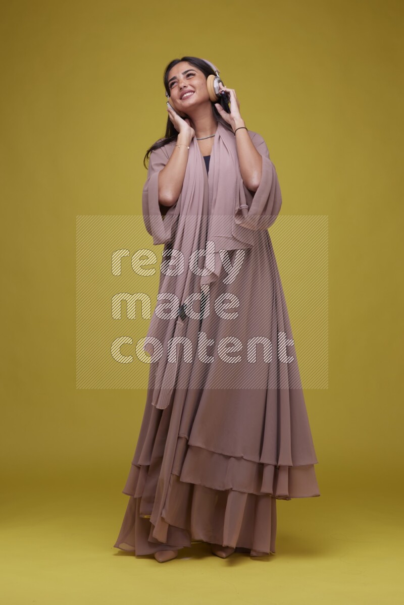 A Saudi woman Listing to Music on a Yellow Background wearing Brown Abaya