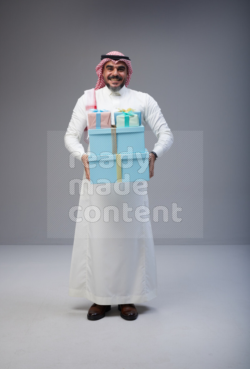 Saudi man Wearing Thob and red Shomag standing holding gift box on Gray background